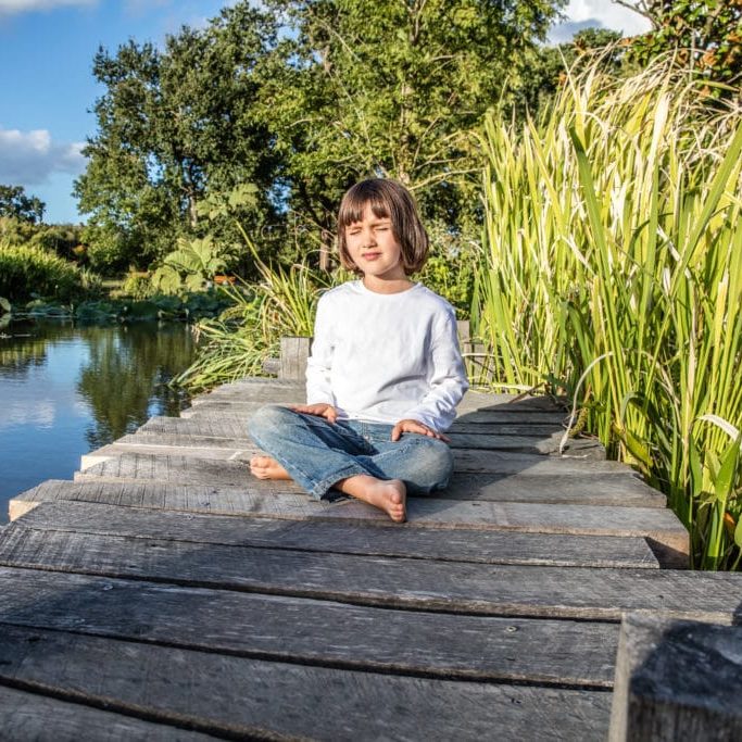 relaxed beautiful young 5-year old child cross-legged with bare feet breathing alone, closing eyes for yoga and meditation near water and trees
